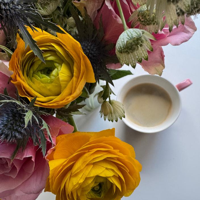 Auf einem Tisch steht eine Tasse Kaffee neben einem Strauß mit Rosen, Ranunkeln und Disteln