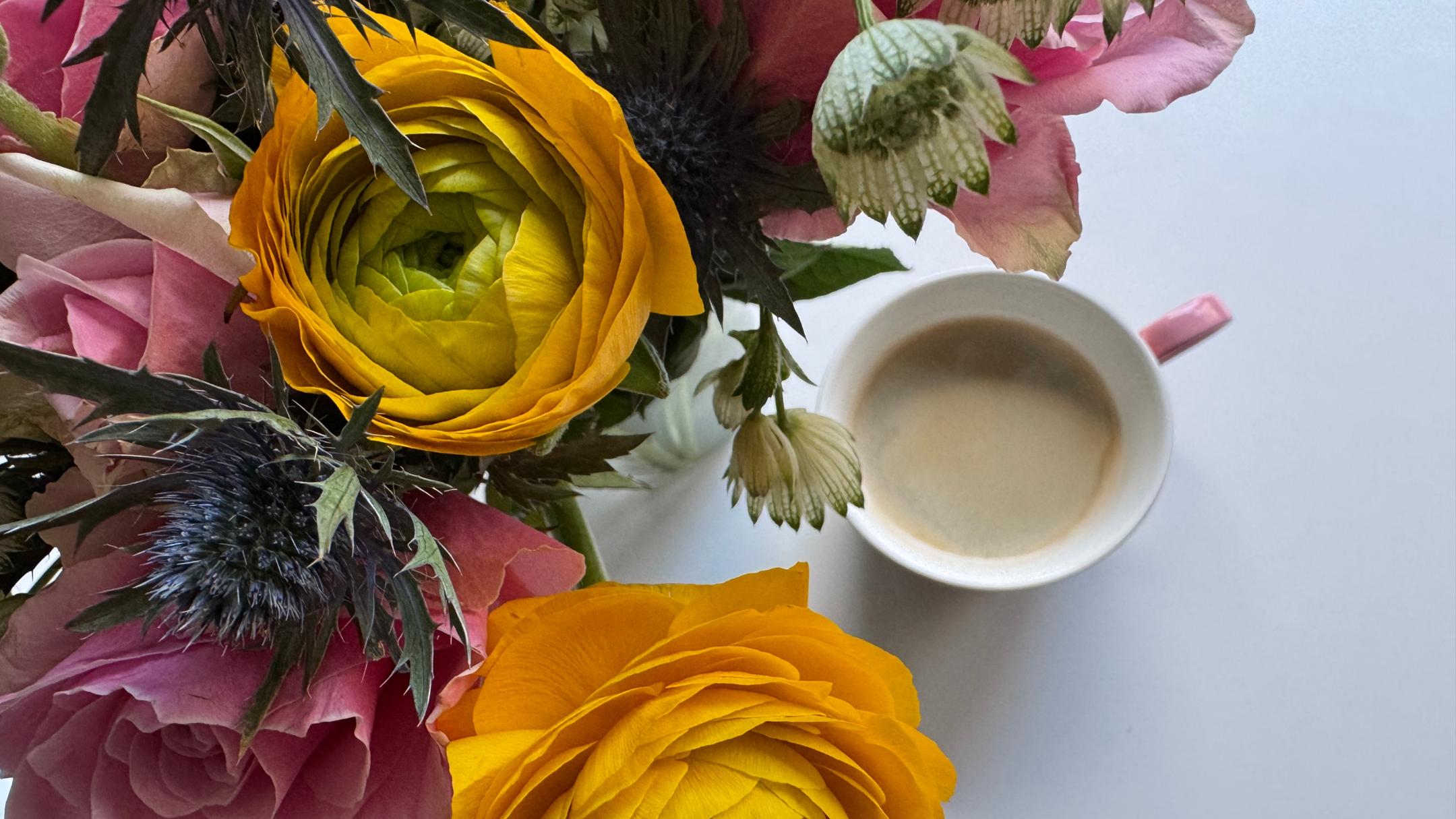 Auf einem Tisch steht eine Tasse Kaffee neben einem Strauß mit Rosen, Ranunkeln und Disteln