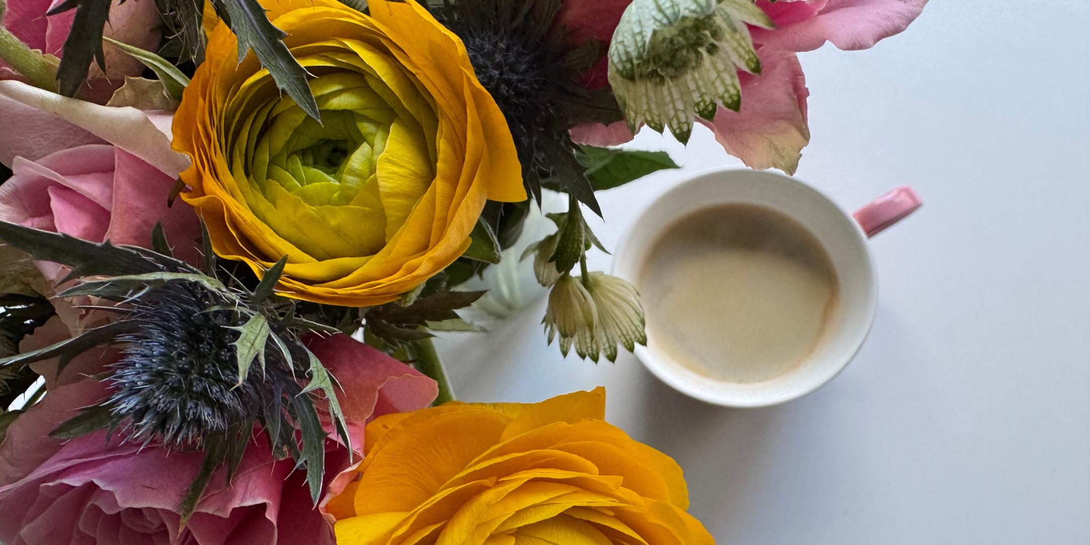 Auf einem Tisch steht eine Tasse Kaffee neben einem Strauß mit Rosen, Ranunkeln und Disteln