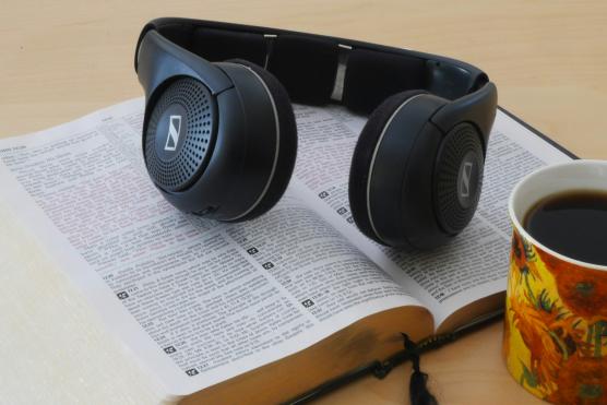 Eine Bibel liegt aufgeschlagen auf dem Tisch. Darauf ein Paar Kopfhörer. Davor stehe eine Tasse mit Kaffee