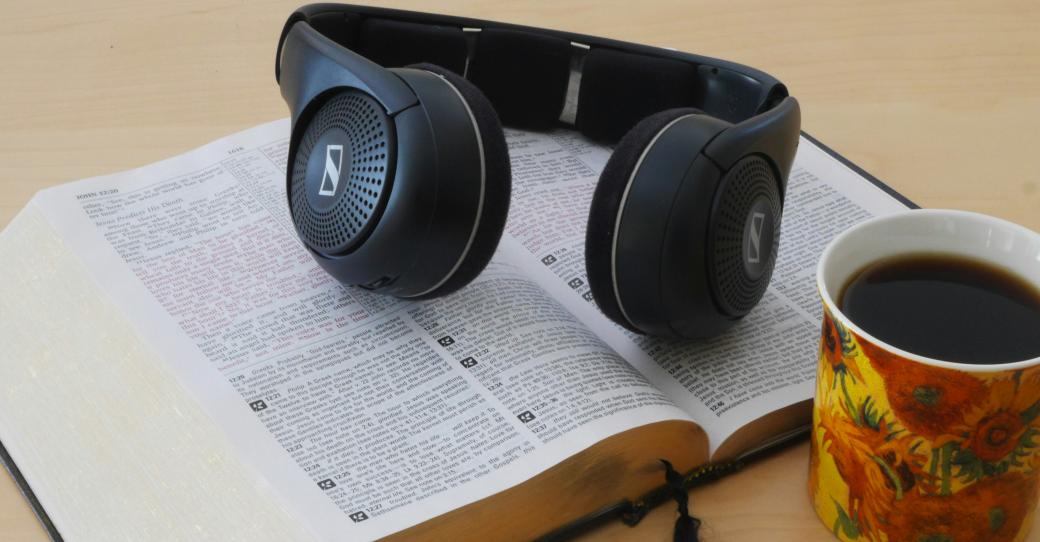 Eine Bibel liegt aufgeschlagen auf dem Tisch. Darauf ein Paar Kopfhörer. Davor stehe eine Tasse mit Kaffee