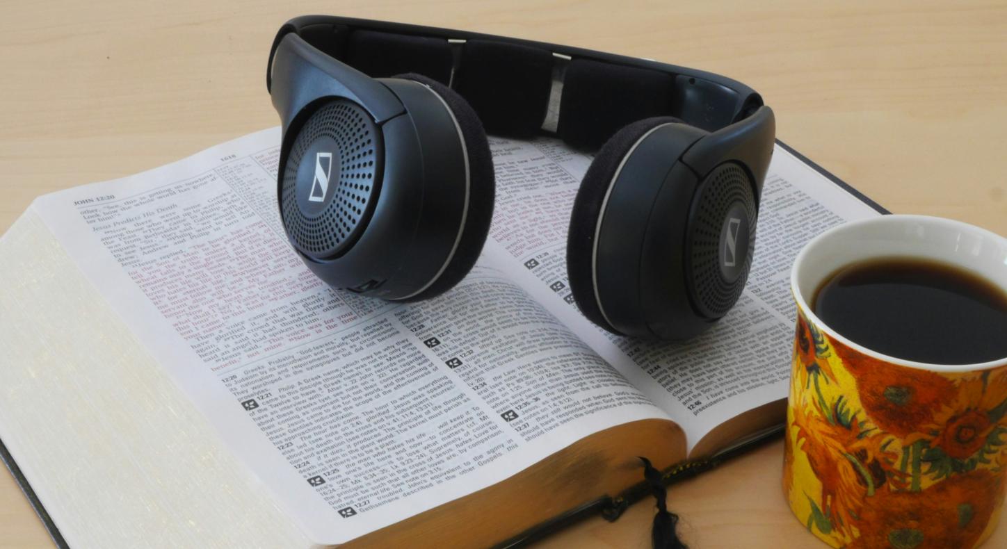 Eine Bibel liegt aufgeschlagen auf dem Tisch. Darauf ein Paar Kopfhörer. Davor stehe eine Tasse mit Kaffee