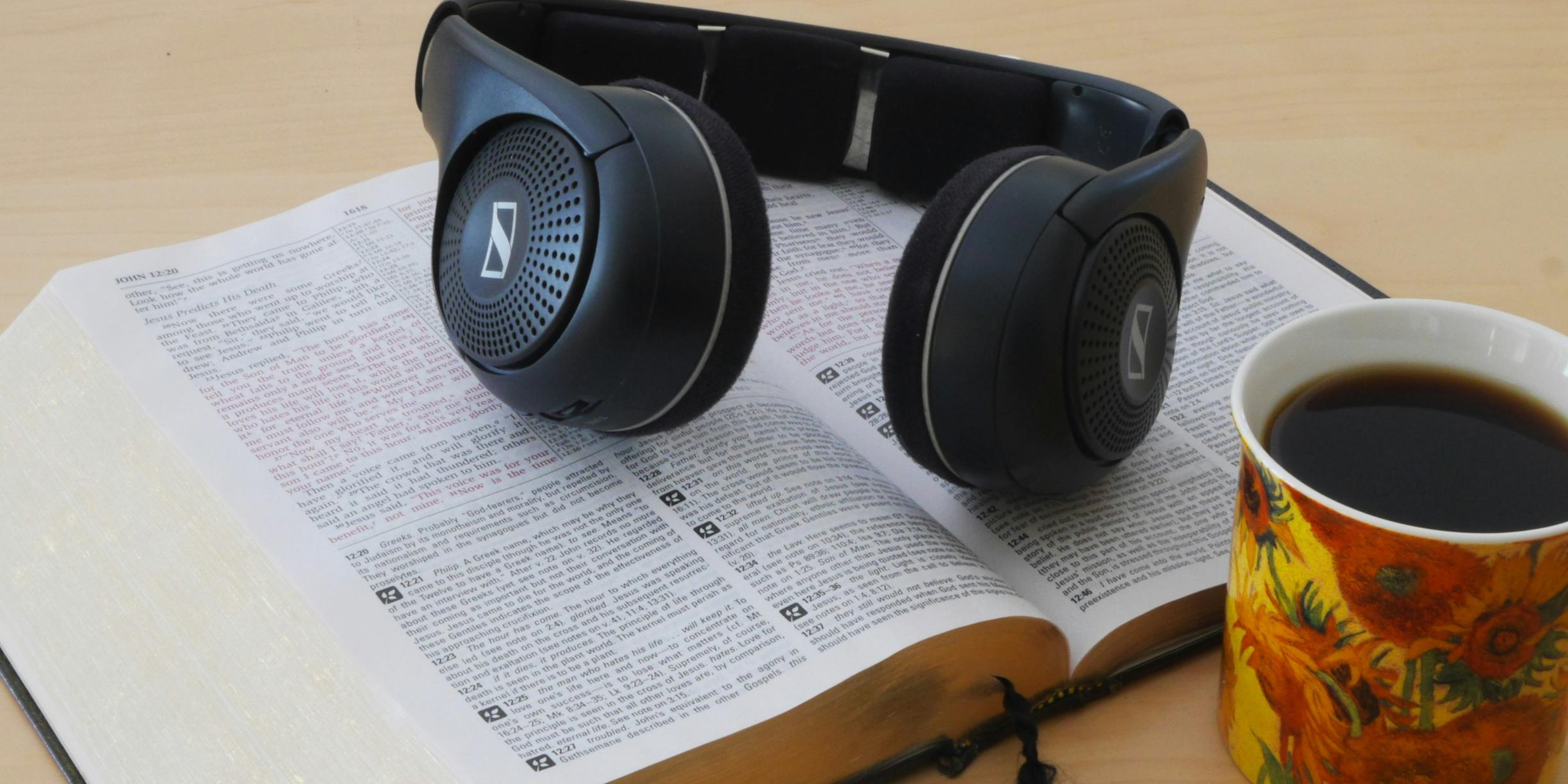 Eine Bibel liegt aufgeschlagen auf dem Tisch. Darauf ein Paar Kopfhörer. Davor stehe eine Tasse mit Kaffee