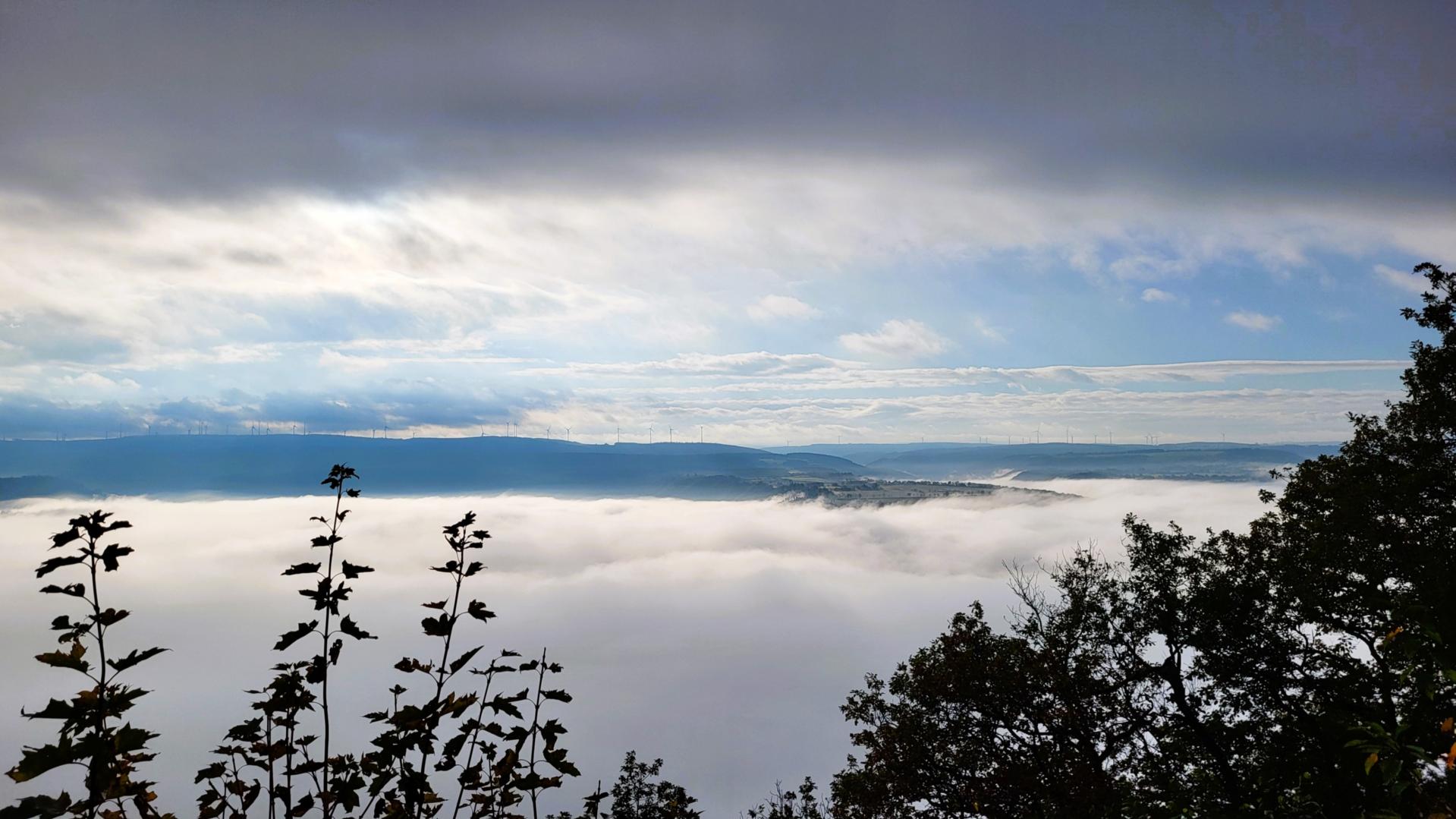 Blick über eine nebelverhangene Landschaft. Im Hintergrund sind Windräder zu sehen. Ein Hügel ragt aus dem Nebel heraus. Der Himmel ist wolkig und sonnig zugleich