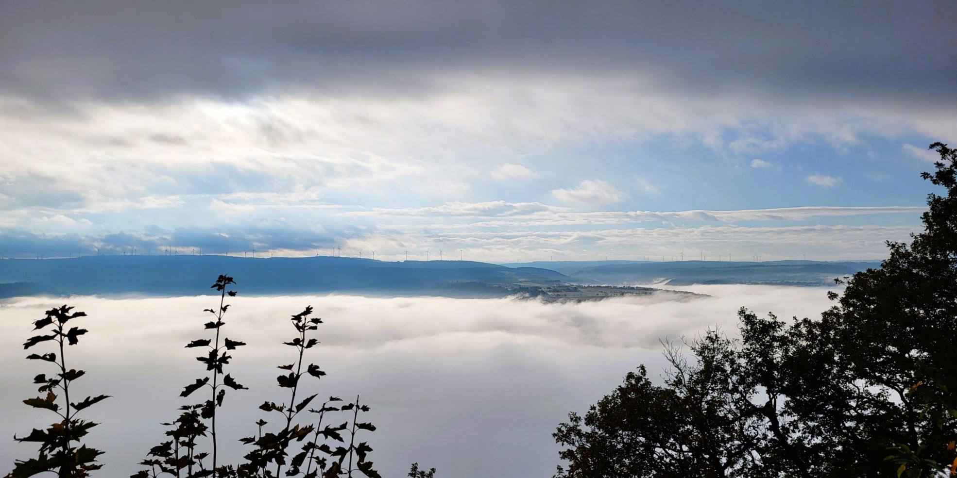 Blick über eine nebelverhangene Landschaft. Im Hintergrund sind Windräder zu sehen. Ein Hügel ragt aus dem Nebel heraus. Der Himmel ist wolkig und sonnig zugleich