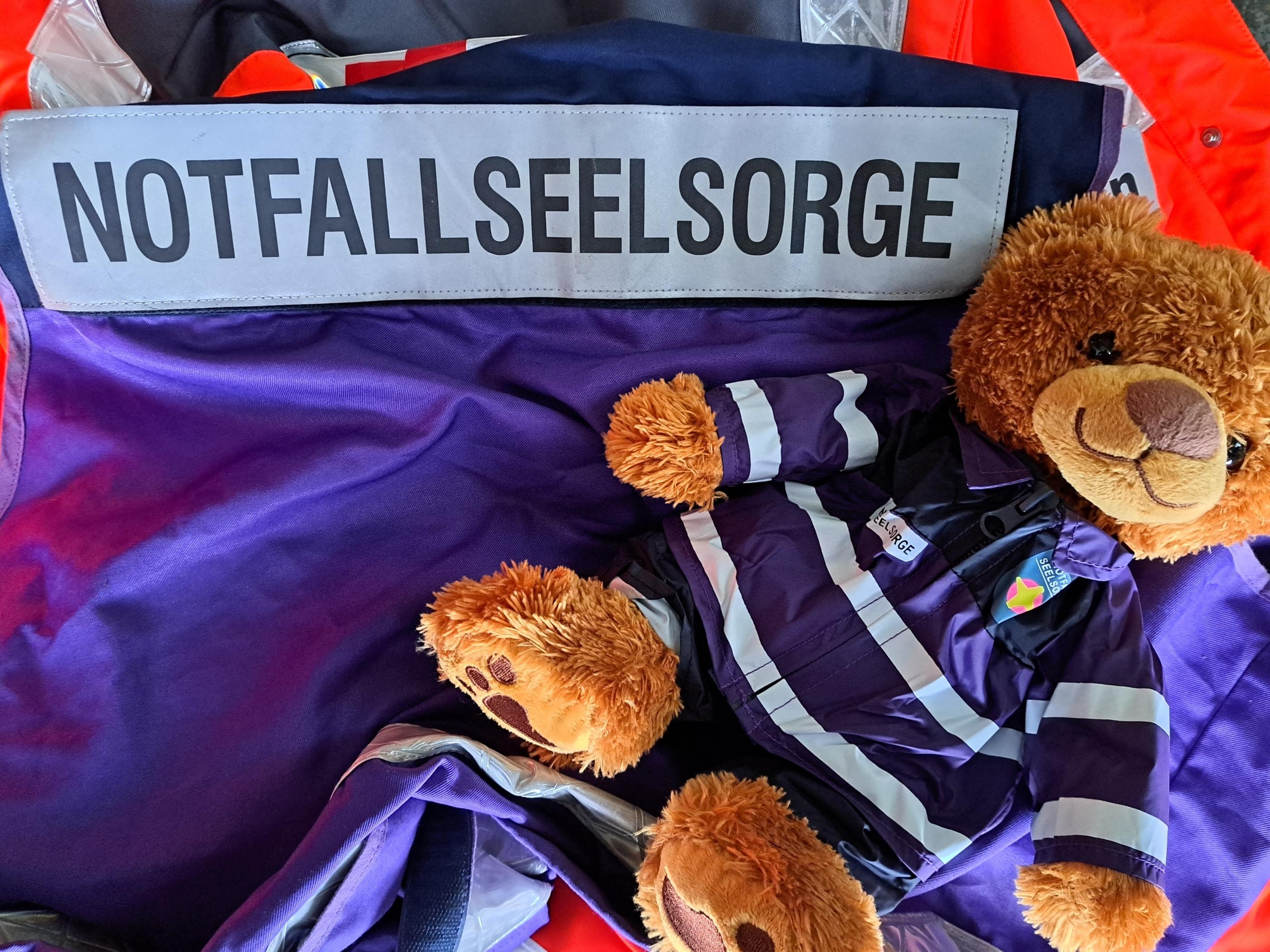 Das Bild zeigt einen braunen Plüschbären in einer lilafarbenen Notfallseelsorge-Uniform mit reflektierenden Streifen. Der Bär liegt auf einer lilafarbenen Stoffunterlage, daneben befindet sich eine Aufschrift 'Notfallseelsorge“. Im Hintergrund sind Teile einer orangenen Warnweste und weitere Utensilien zu sehen.