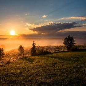 Sonnenaufgang über einer hügeligen grünen Wiese; Nebel liegt auf den Wiesen; die Sonne ist rötlich, der Himmel wird langsam Blau.
