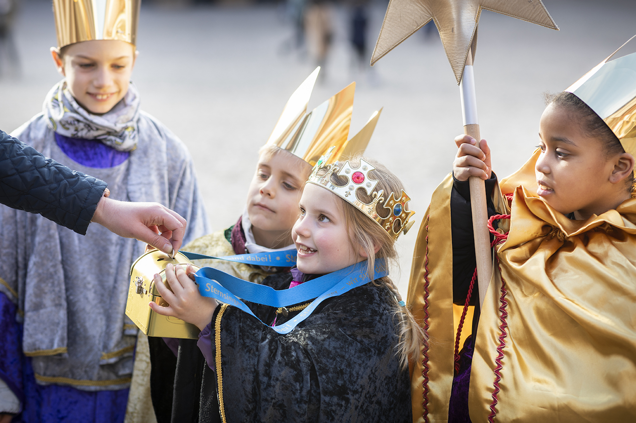 als Sternsinger verkleidete Kinder halten eine Spendendose hin; eine Hand wirf Geld hinein