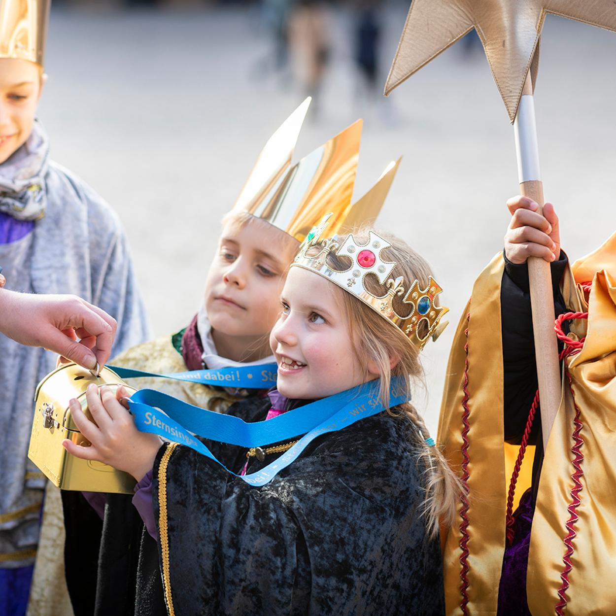 als Sternsinger verkleidete Kinder halten eine Spendendose hin; eine Hand wirf Geld hinein