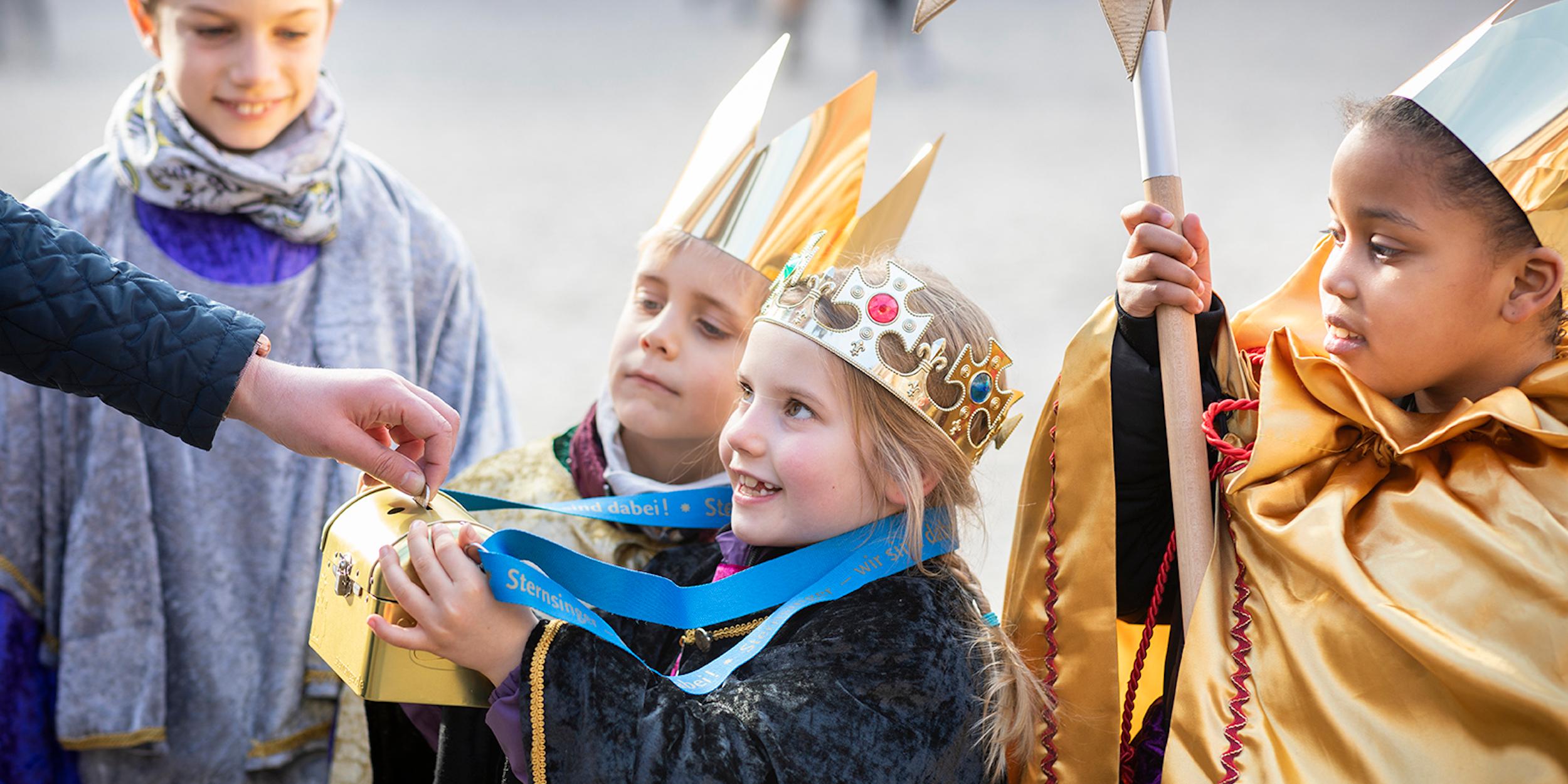 als Sternsinger verkleidete Kinder halten eine Spendendose hin; eine Hand wirf Geld hinein