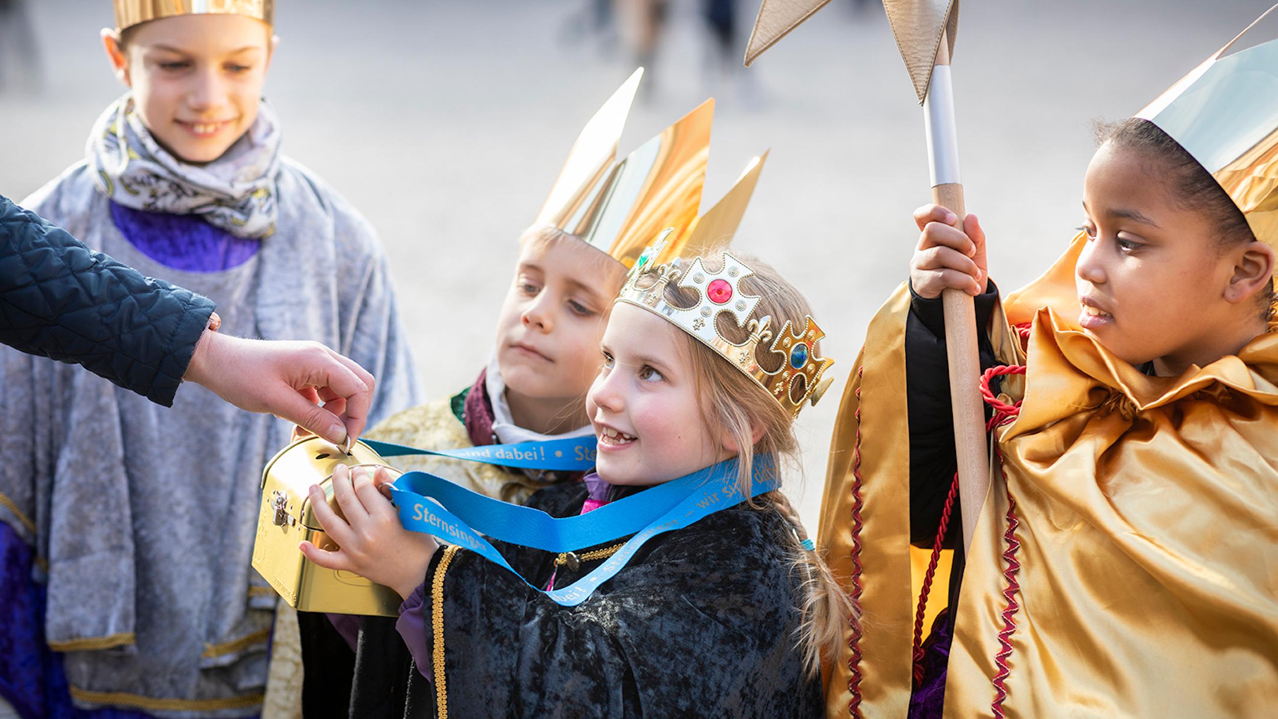 als Sternsinger verkleidete Kinder halten eine Spendendose hin; eine Hand wirf Geld hinein