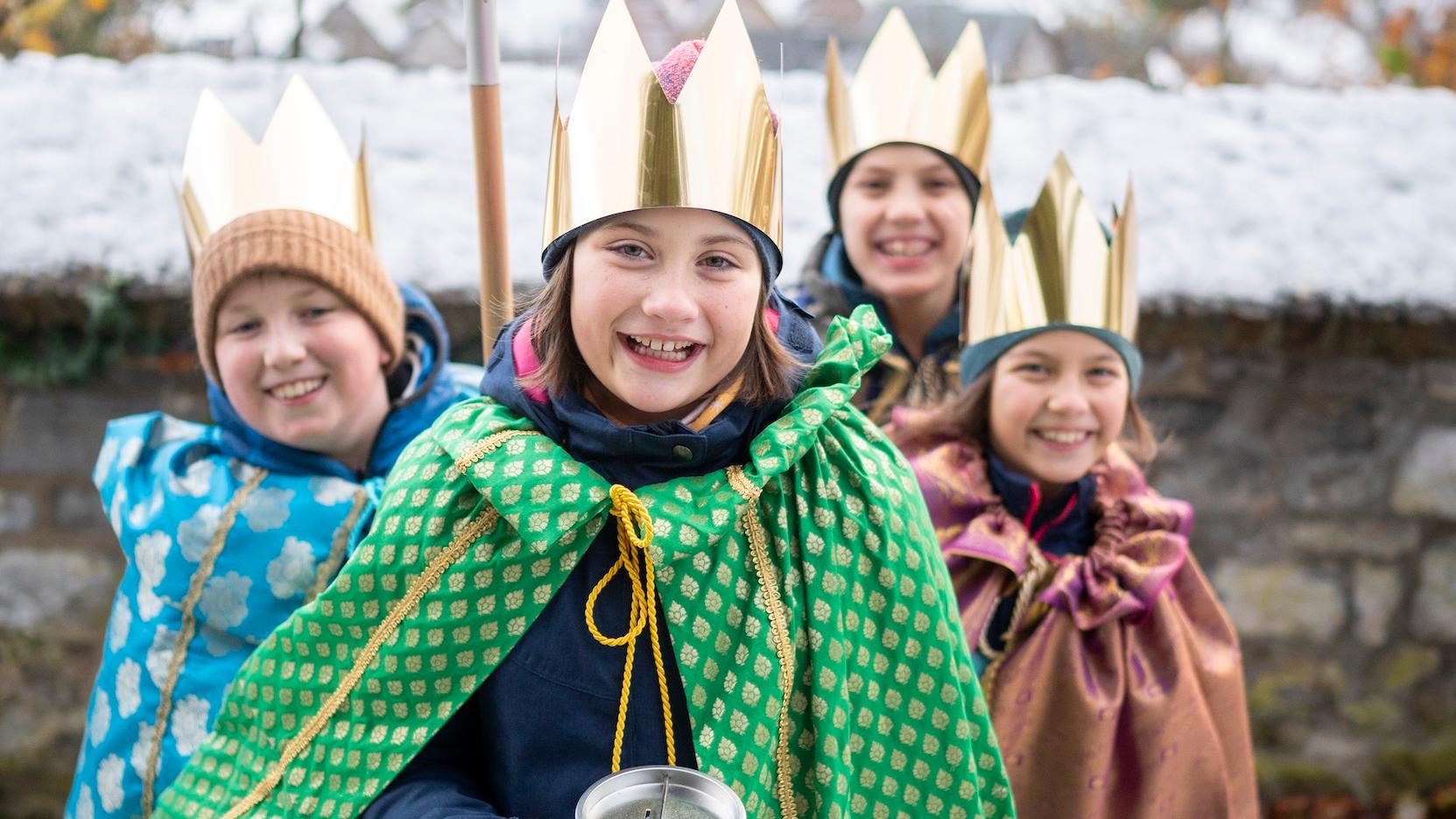 als Sternsinger verkleidete Kinder stehen als Gruppe zusammen, lächeln; einer hält einen Stern hoch, der vordere hält eine Spendendose
