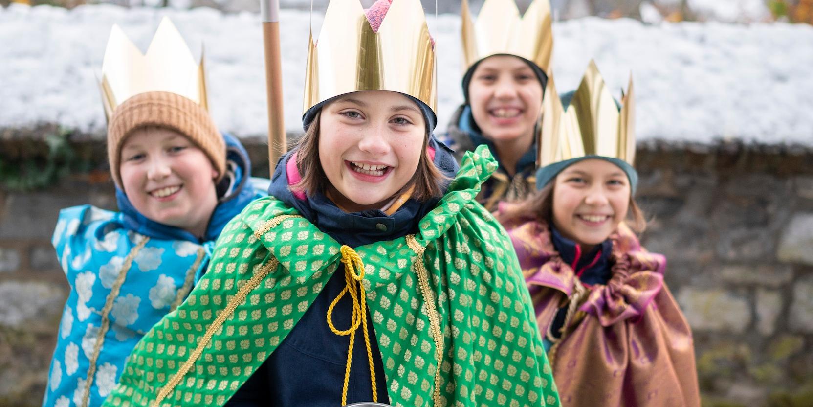 Sternsinger als Sternsinger verkleidete Kinder stehen als Gruppe zusammen, lächeln; einer hält einen Stern hoch, der vordere hält eine Spendendose