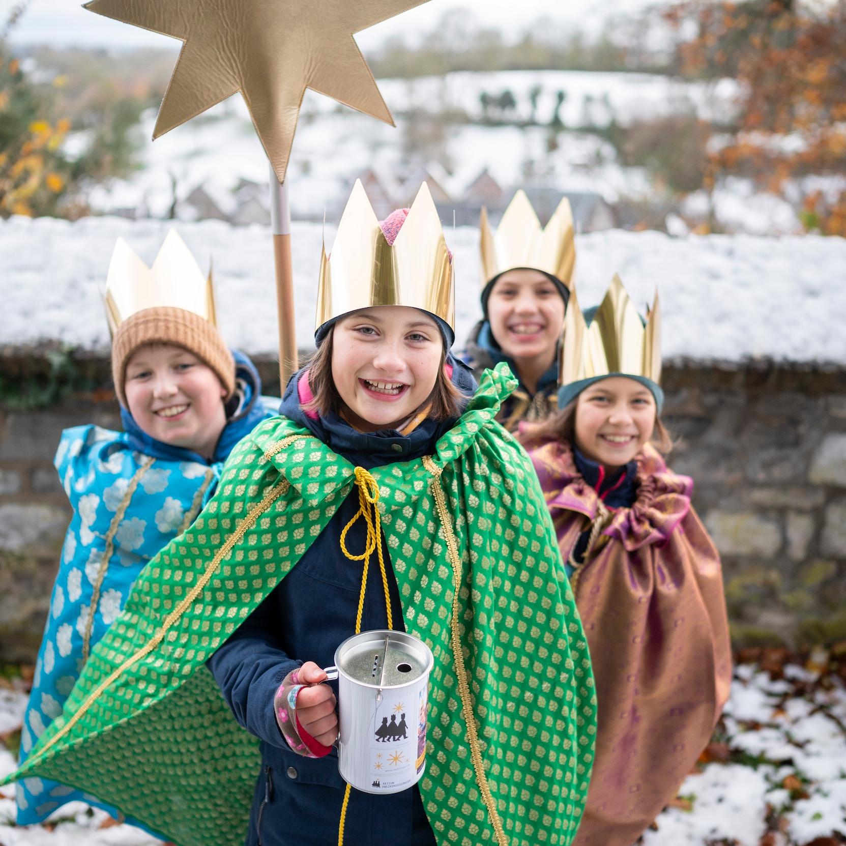 als Sternsinger verkleidete Kinder stehen als Gruppe zusammen, lächeln; einer hält einen Stern hoch, der vordere hält eine Spendendose
