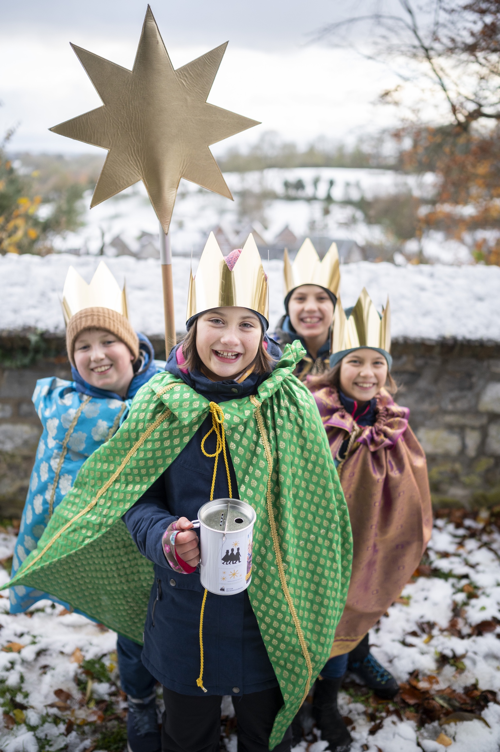 als Sternsinger verkleidete Kinder stehen als Gruppe zusammen, lächeln; einer hält einen Stern hoch, der vordere hält eine Spendendose