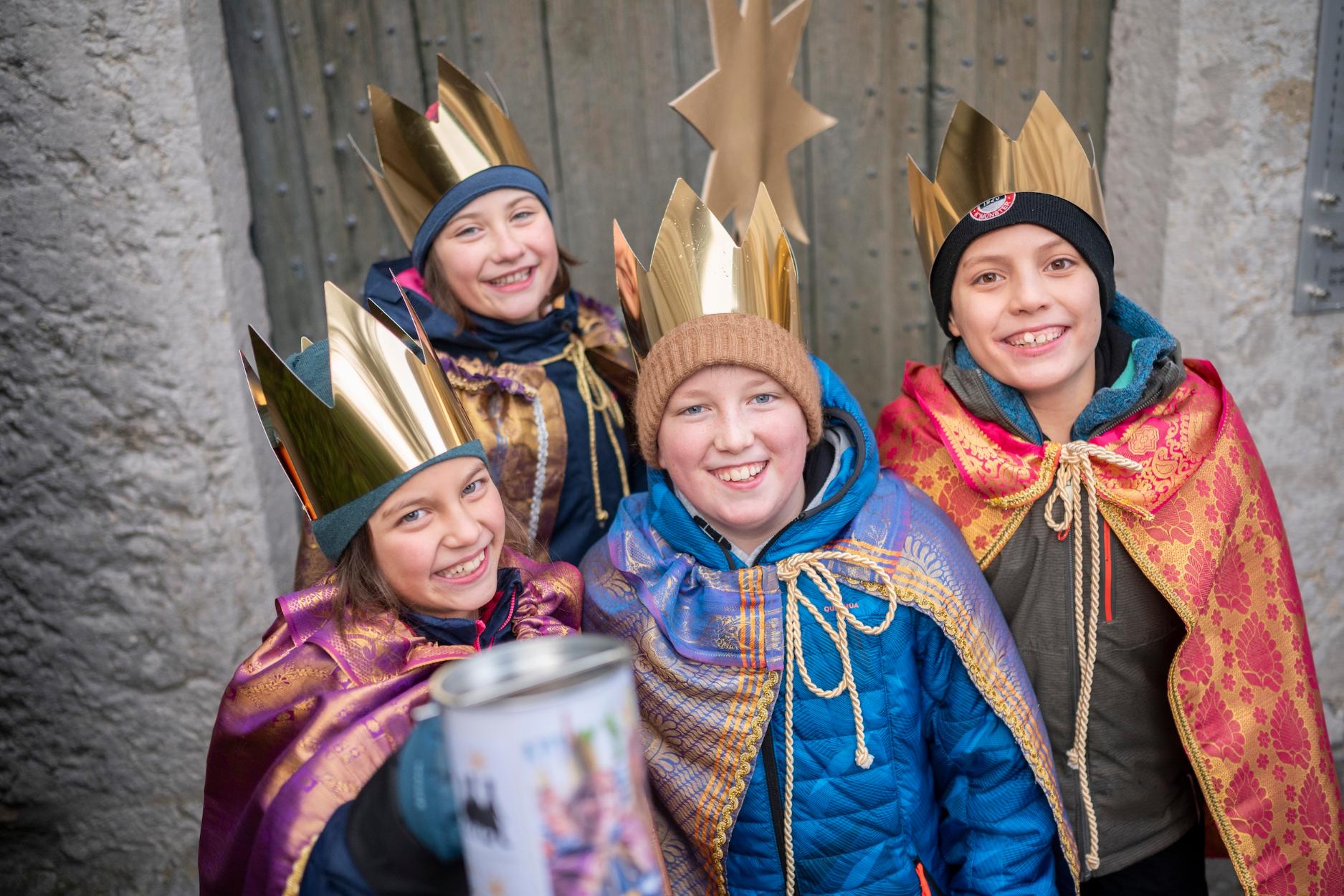 Vier als Sternsinger verkleidete Kinder mit Stern lächeln und halten eine Spendendose in die Kamera