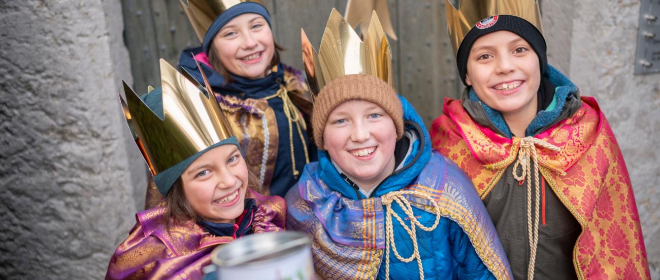 Vier als Sternsinger verkleidete Kinder mit Stern lächeln und halten eine Spendendose in die Kamera