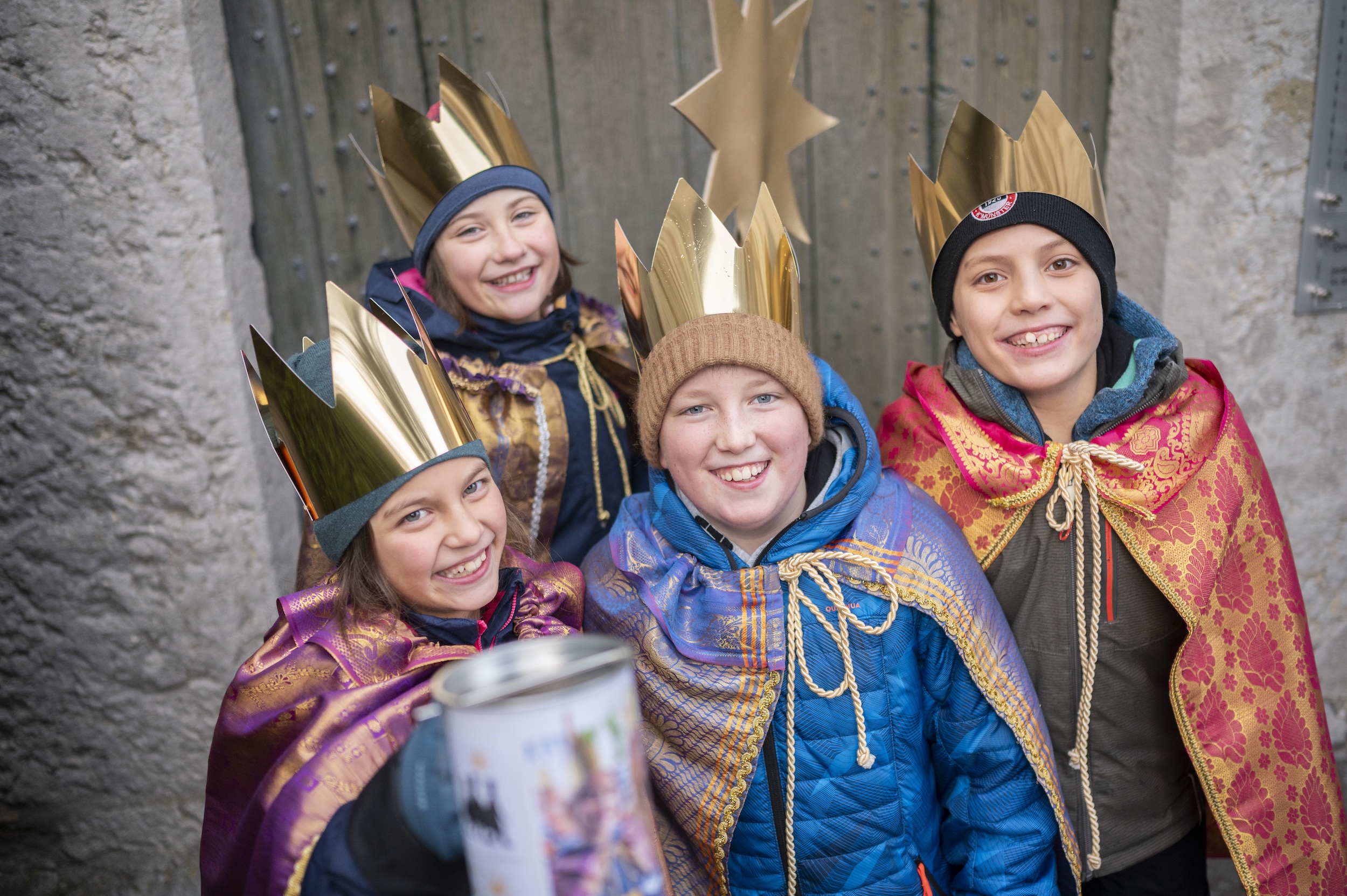 Vier als Sternsinger verkleidete Kinder mit Stern lächeln und halten eine Spendendose in die Kamera
