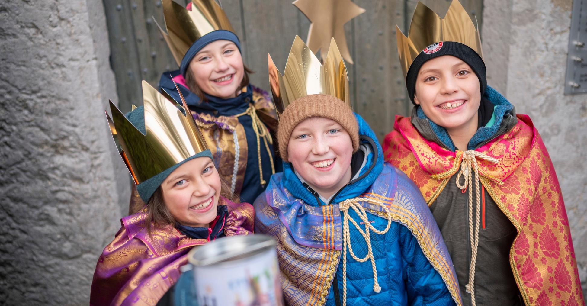Vier als Sternsinger verkleidete Kinder mit Stern lächeln und halten eine Spendendose in die Kamera