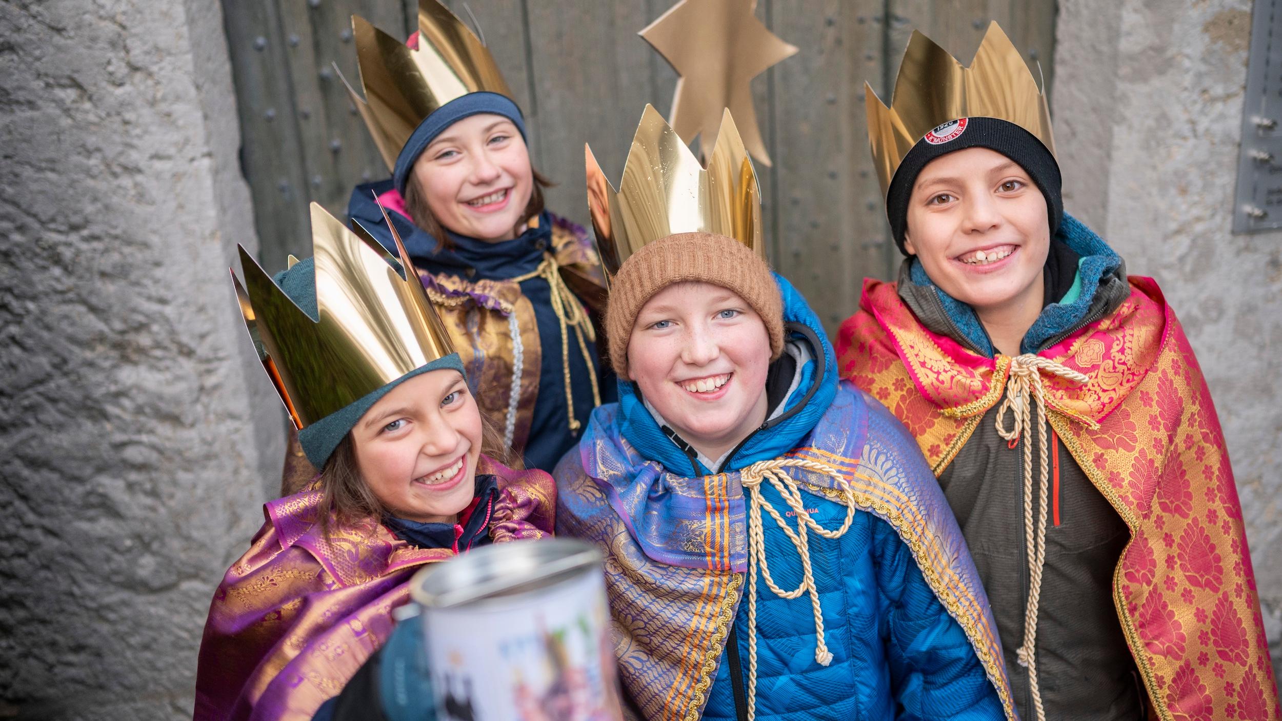 Vier als Sternsinger verkleidete Kinder mit Stern lächeln und halten eine Spendendose in die Kamera