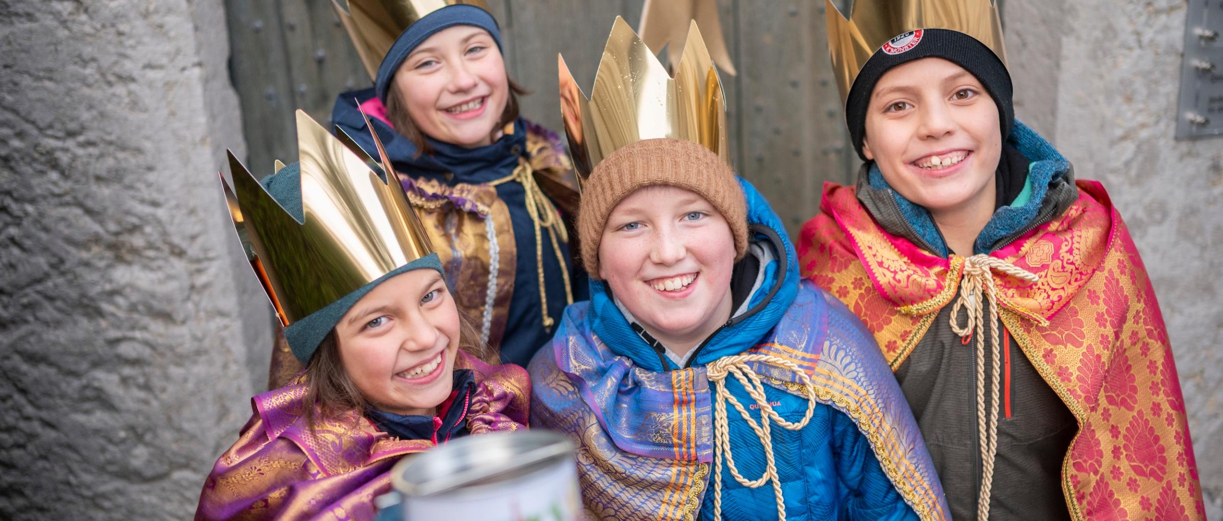 Vier als Sternsinger verkleidete Kinder mit Stern lächeln und halten eine Spendendose in die Kamera