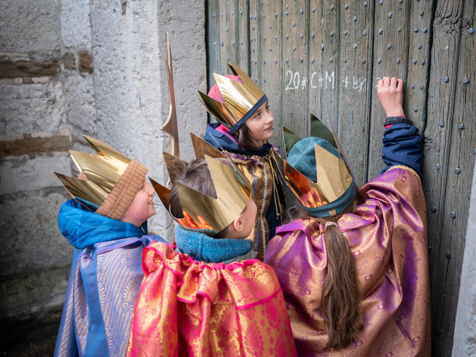 als Sternsinger verkleidete Kinder schreiben mit Kreide einen Segen an eine Holztür