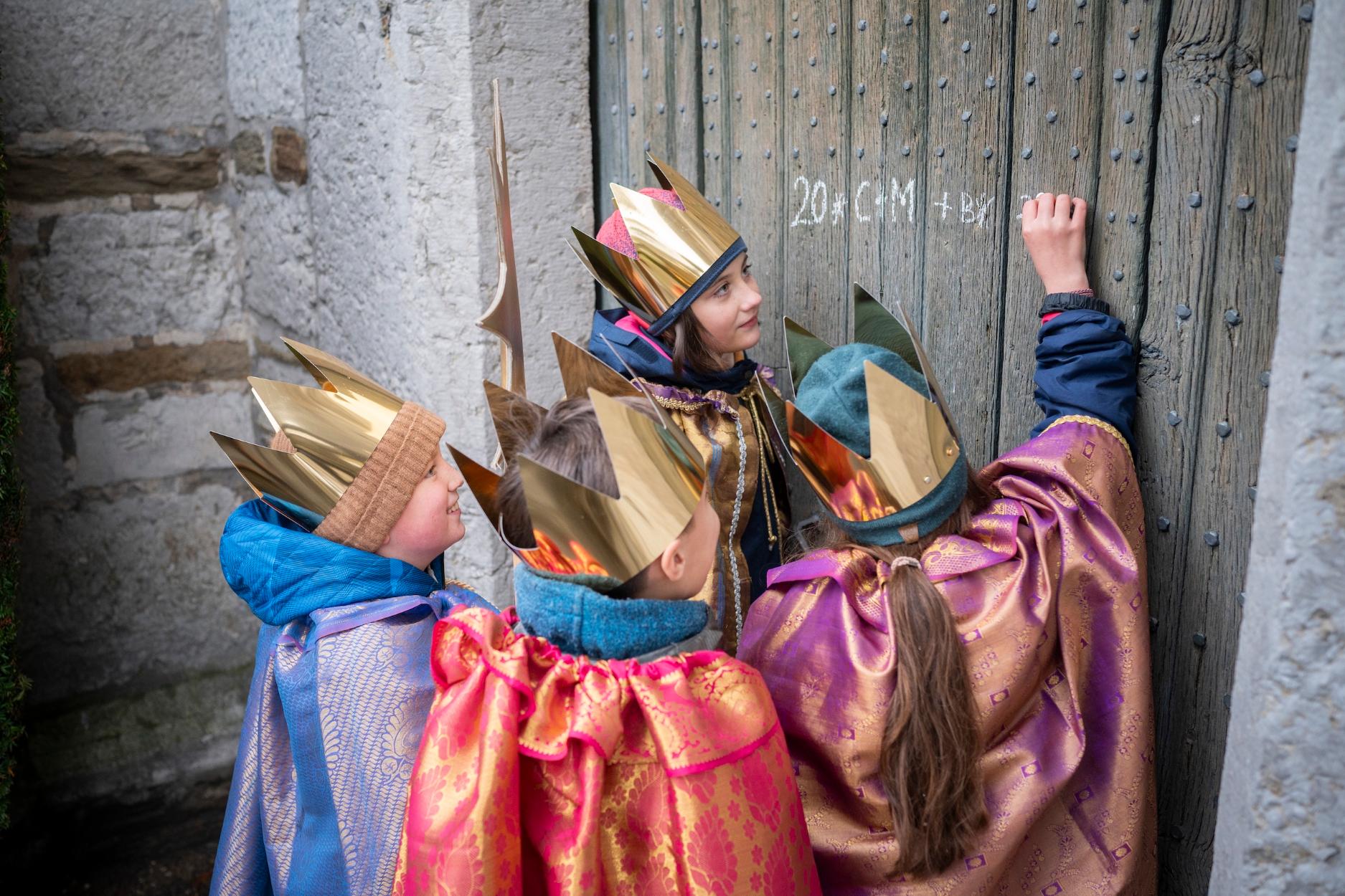 als Sternsinger verkleidete Kinder schreiben mit Kreide einen Segen an eine Holztür