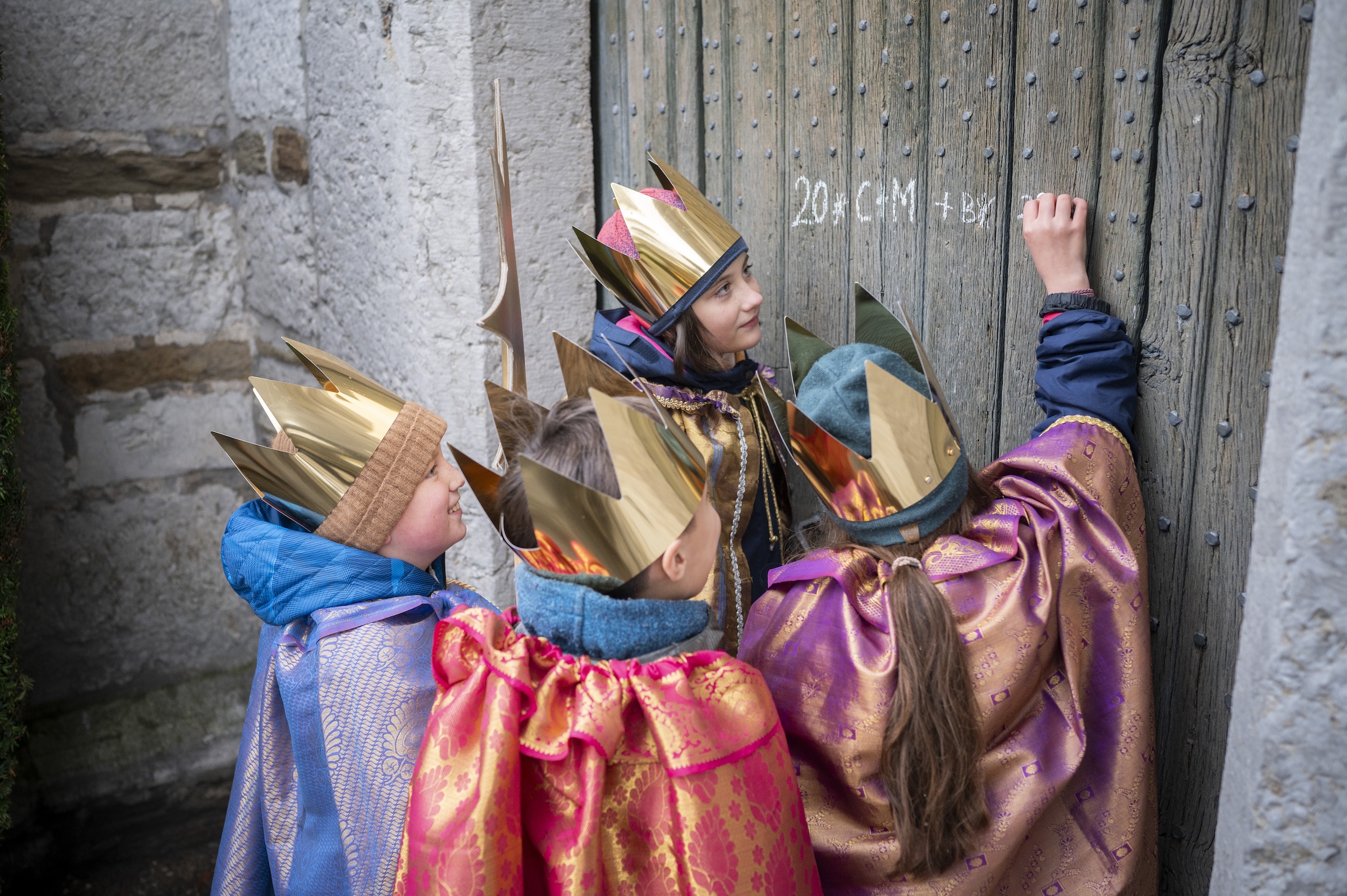 Sternsinger (c) Mika Vaeisaenen/Kindermissionswerk als Sternsinger verkleidete Kinder schreiben mit Kreide einen Segen an eine Holztür