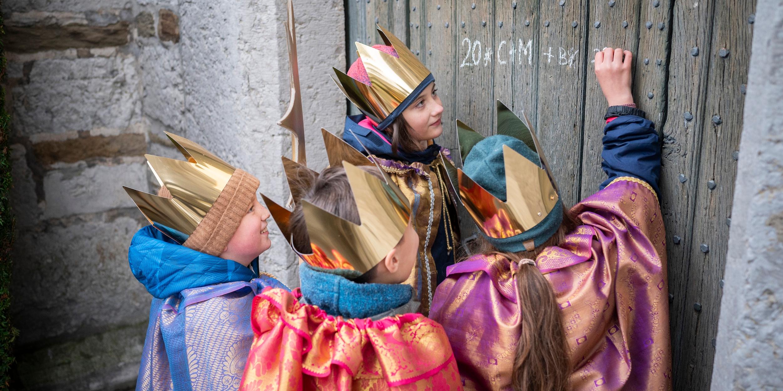 als Sternsinger verkleidete Kinder schreiben mit Kreide einen Segen an eine Holztür