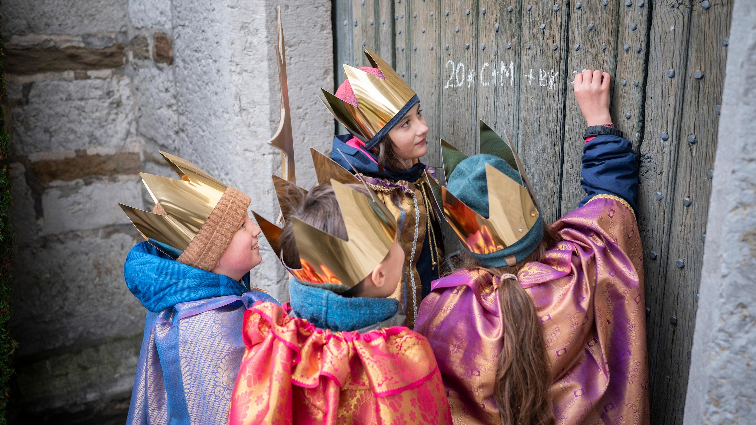 als Sternsinger verkleidete Kinder schreiben mit Kreide einen Segen an eine Holztür