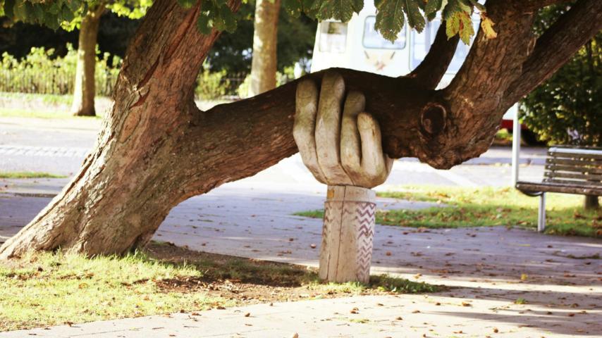 Eine große Hand aus Holz stützt einen Baumstamm, der sich zur Seite geneigt hat