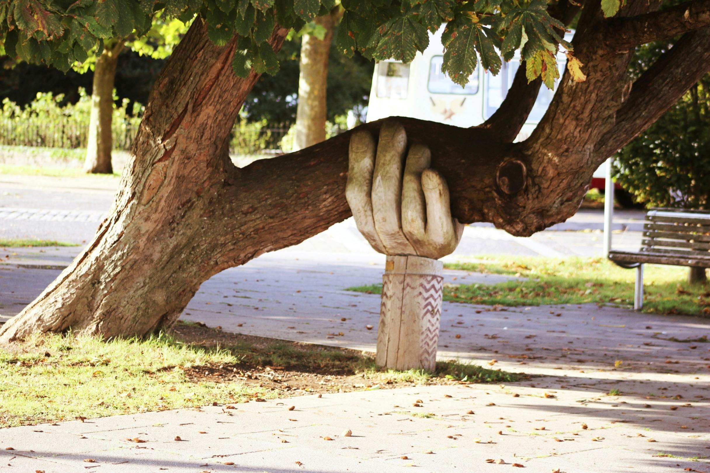 Eine große Hand aus Holz stützt einen Baumstamm, der sich zur Seite geneigt hat