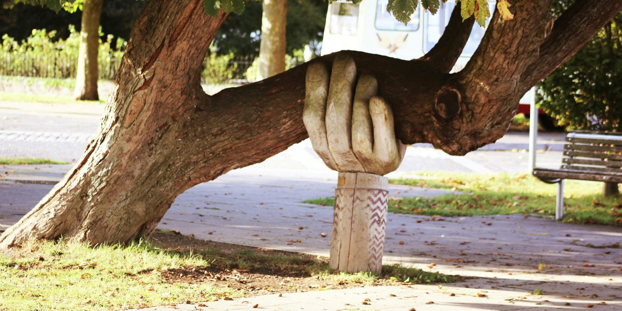 Eine große Hand aus Holz stützt einen Baumstamm, der sich zur Seite geneigt hat