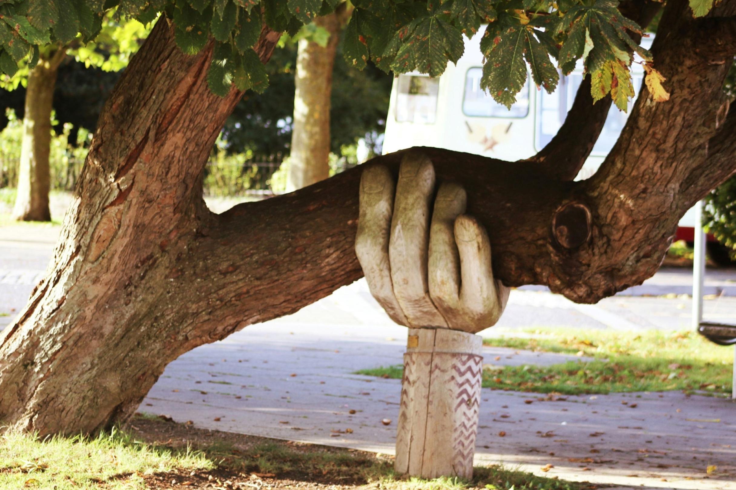 Eine große Hand aus Holz stützt einen Baumstamm, der sich zur Seite geneigt hat