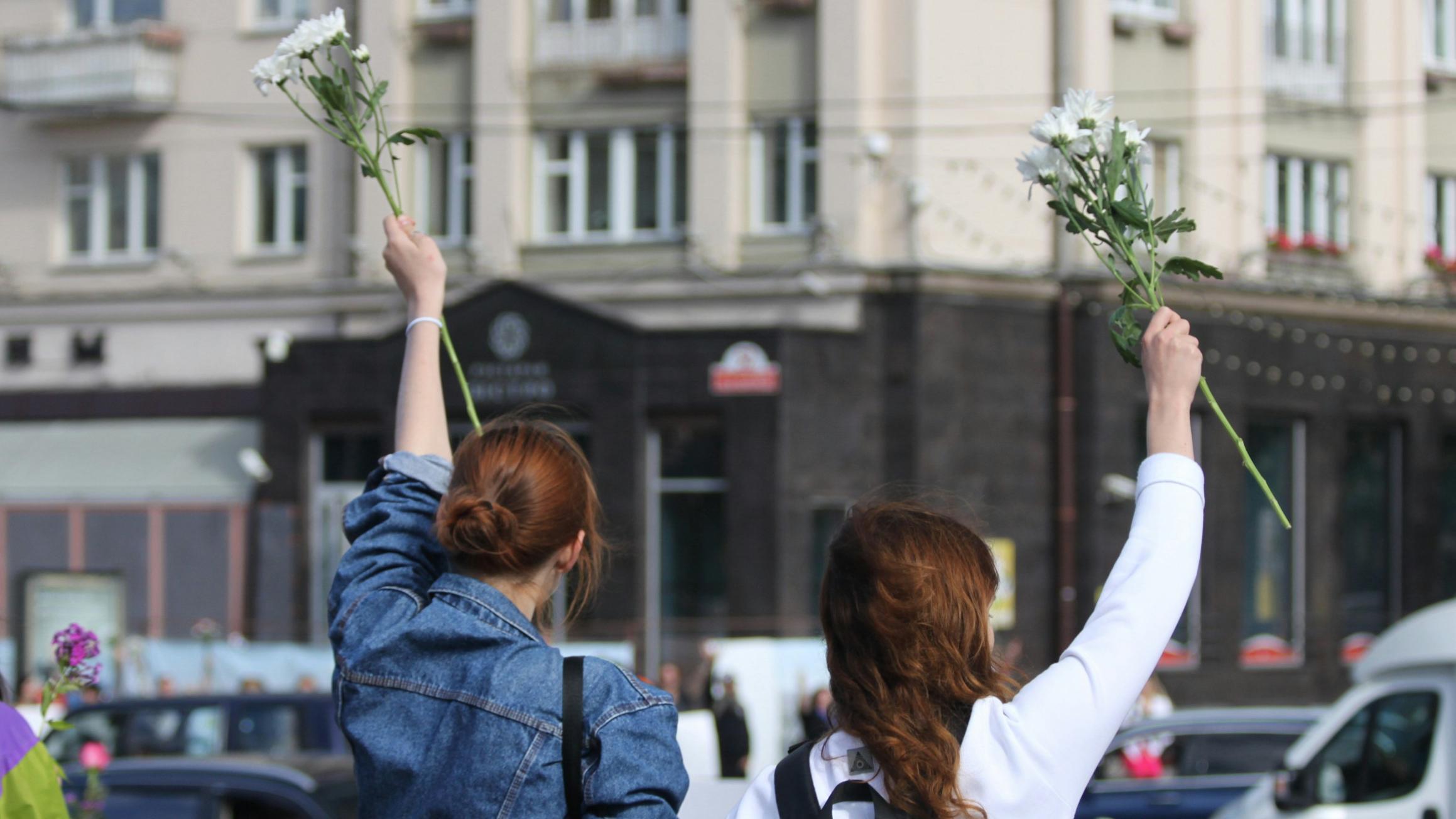 Zwei Frauen, von hinten gesehen, stehen an einer Straße und halten Blumen in die Luft.