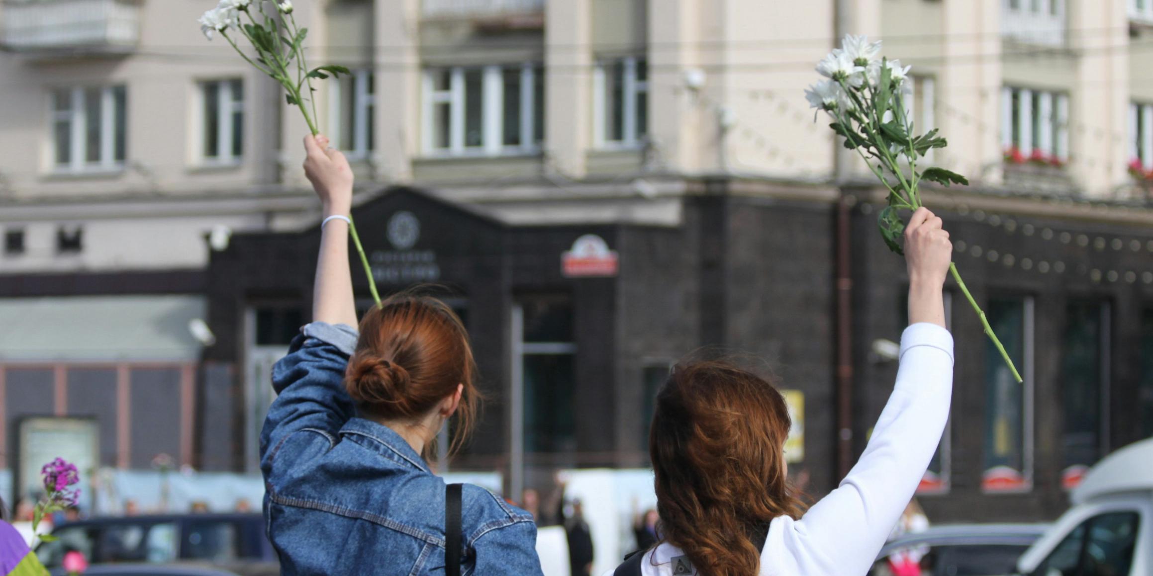 Symbolbild (c) Alesia/unsplash.com Zwei Frauen, von hinten gesehen, stehen an einer Straße und halten Blumen in die Luft.