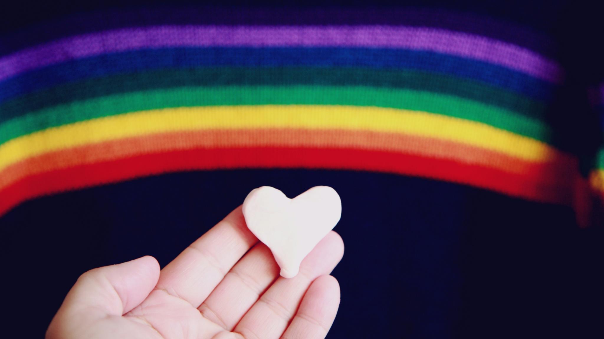 Eine Hand hält ein Herz aus Knete. Im Hintergrund ist ein Regenbogen in den Farben lila, blau, grün, gelb, orange und rot