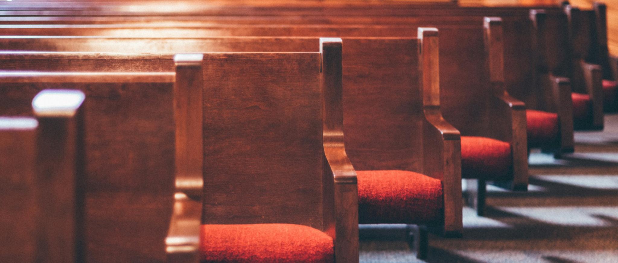 Man sieht einige Reihen von Kirchenbänken aus Holz mit roten Sitzpolstern