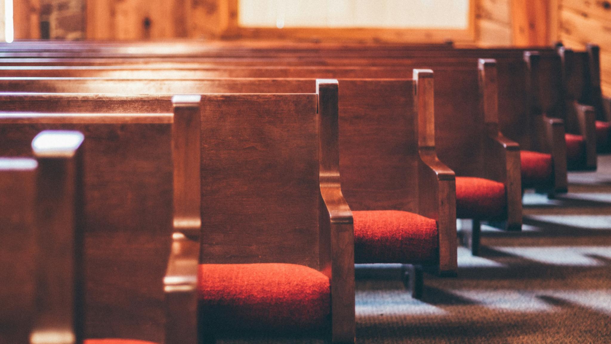 Man sieht einige Reihen von Kirchenbänken aus Holz mit roten Sitzpolstern