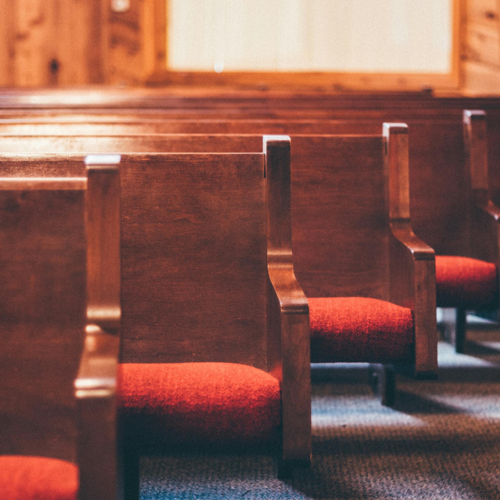 Man sieht einige Reihen von Kirchenbänken aus Holz mit roten Sitzpolstern