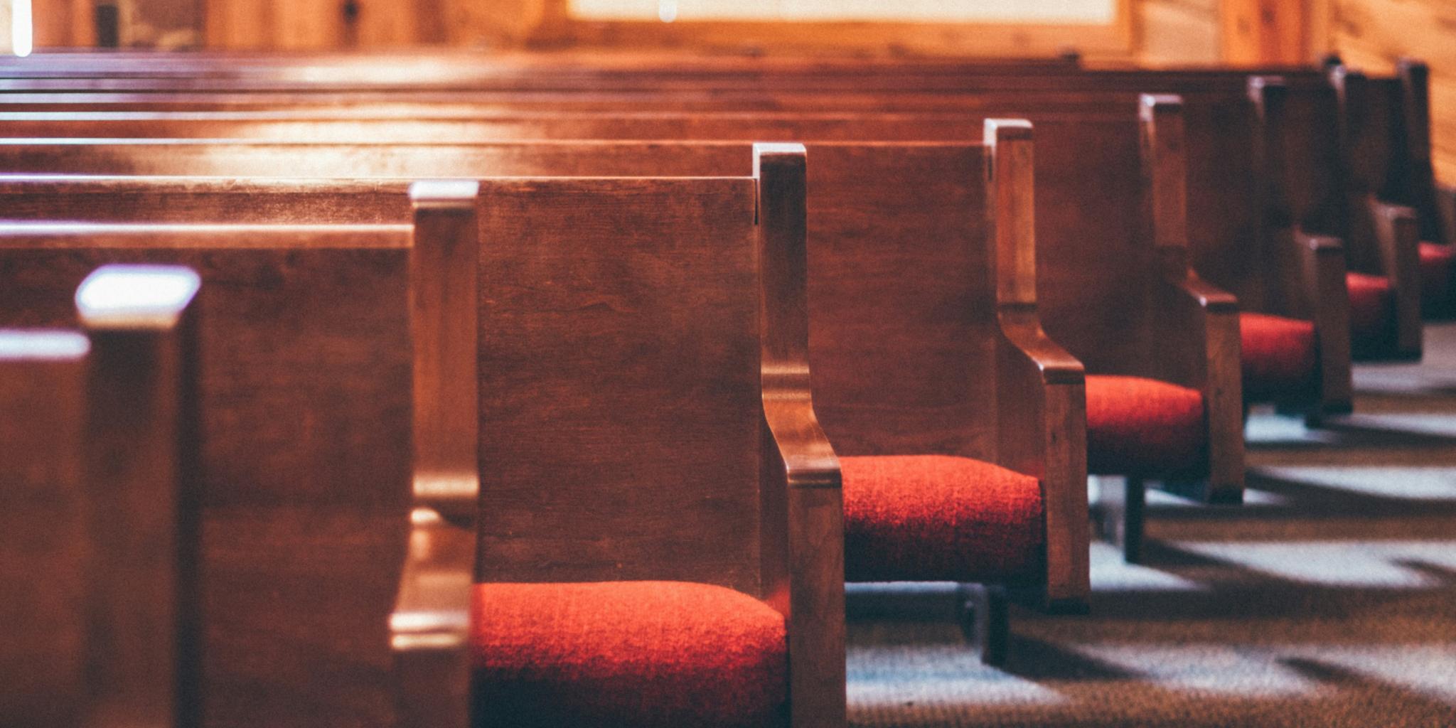 Man sieht einige Reihen von Kirchenbänken aus Holz mit roten Sitzpolstern