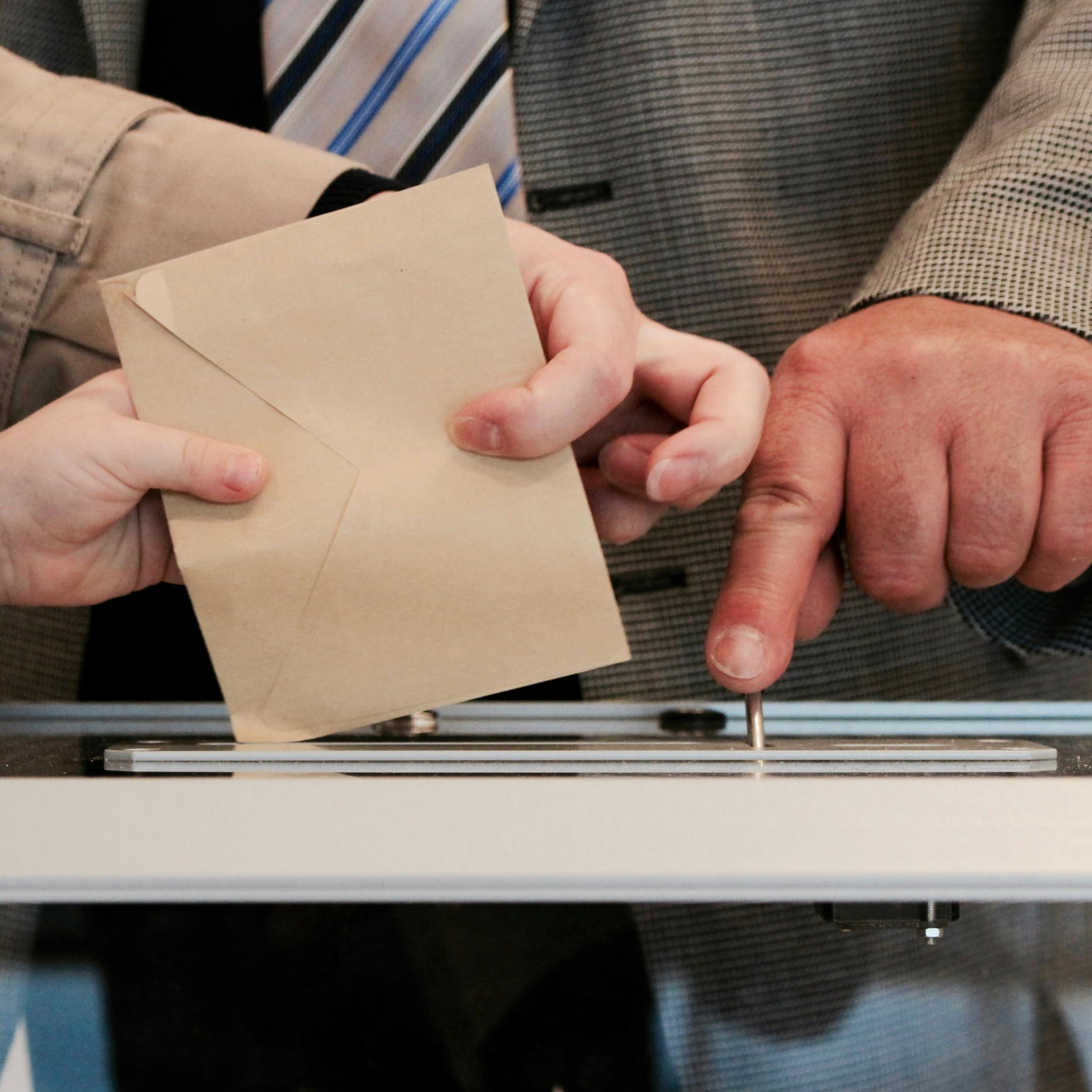 Hände werfen einen Umschlag in eine Wahlurne