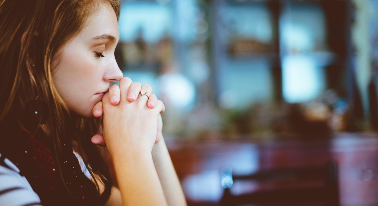 Eine Frau sitzt an einem Tischen, die Ellbogen abgestützt und die Hände zum Gebet gefaltet
