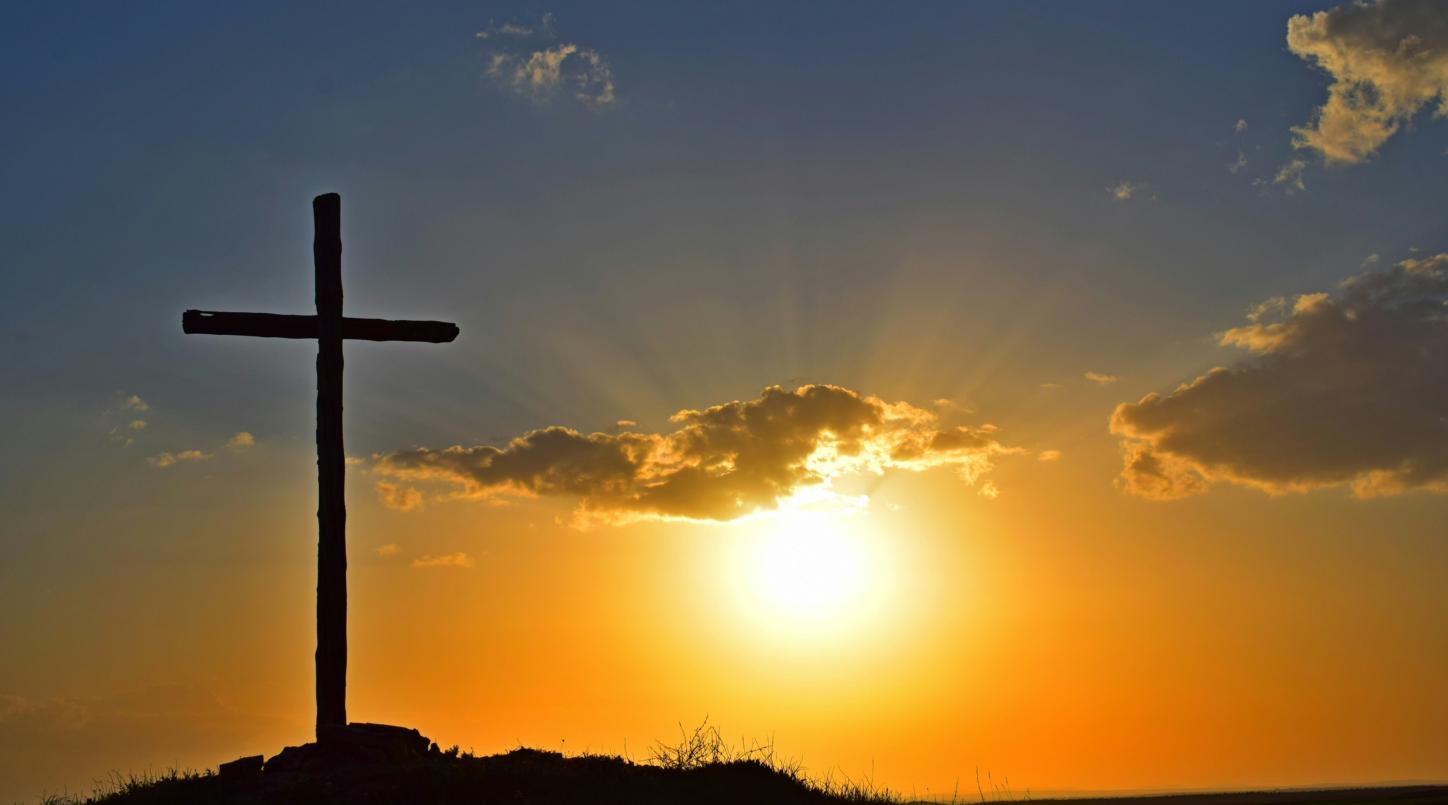 Auf einem Berg steht ein großes Kreuz, dahinter geht die Sonne unter