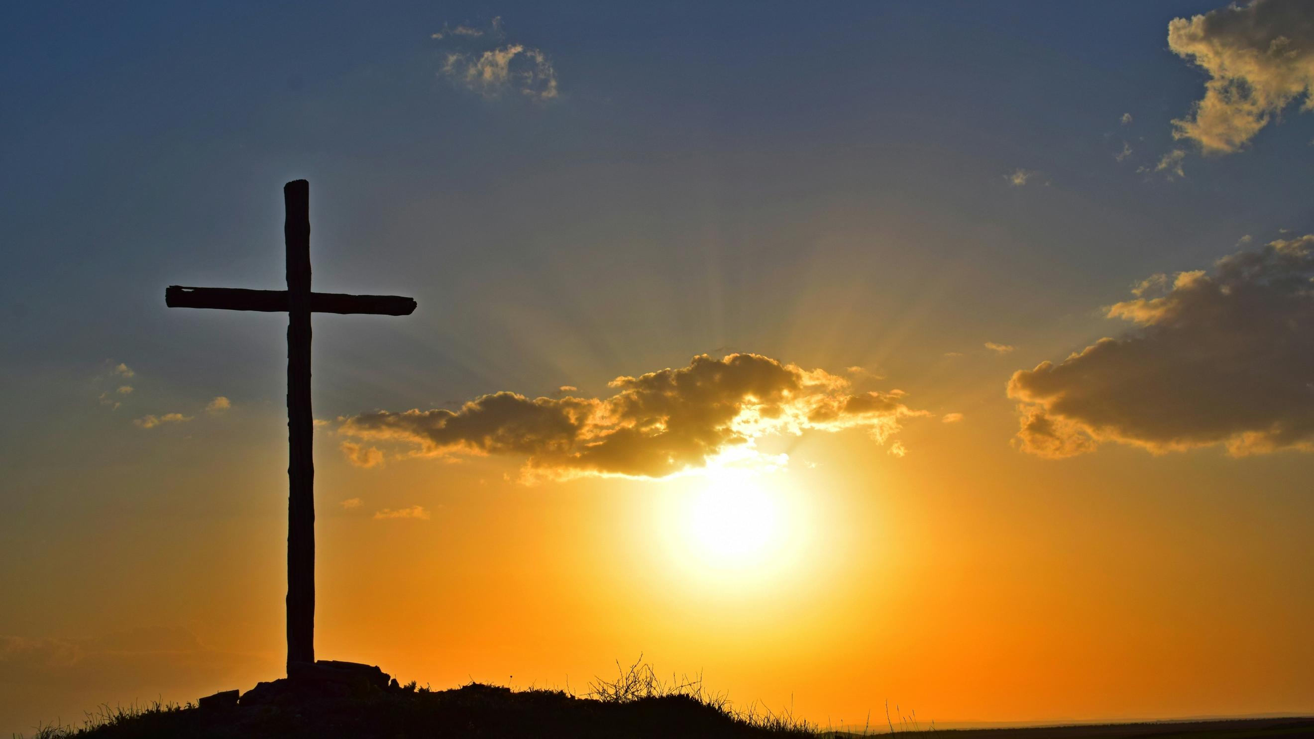 Auf einem Berg steht ein großes Kreuz, dahinter geht die Sonne unter
