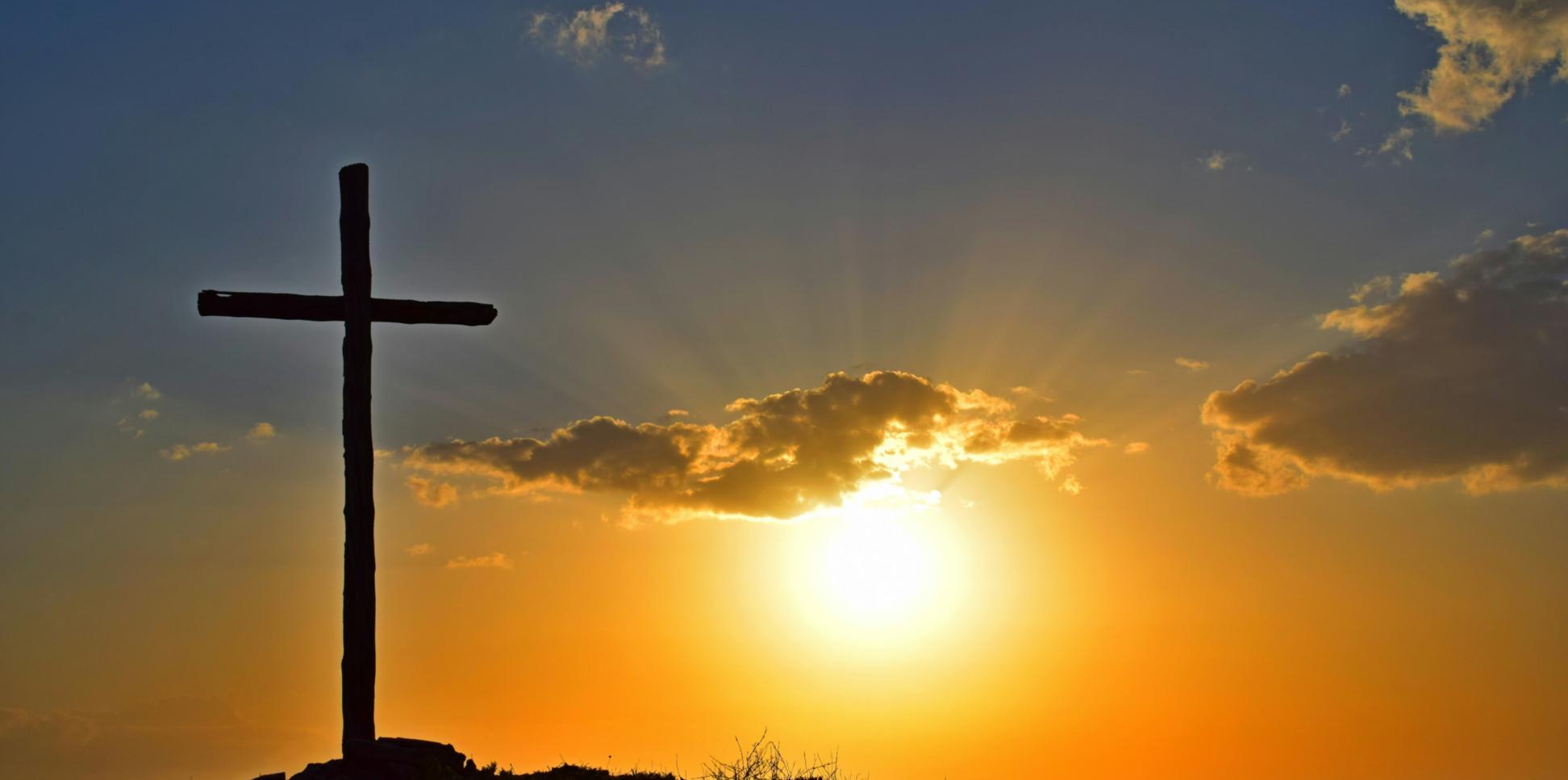 Auf einem Berg steht ein großes Kreuz, dahinter geht die Sonne unter