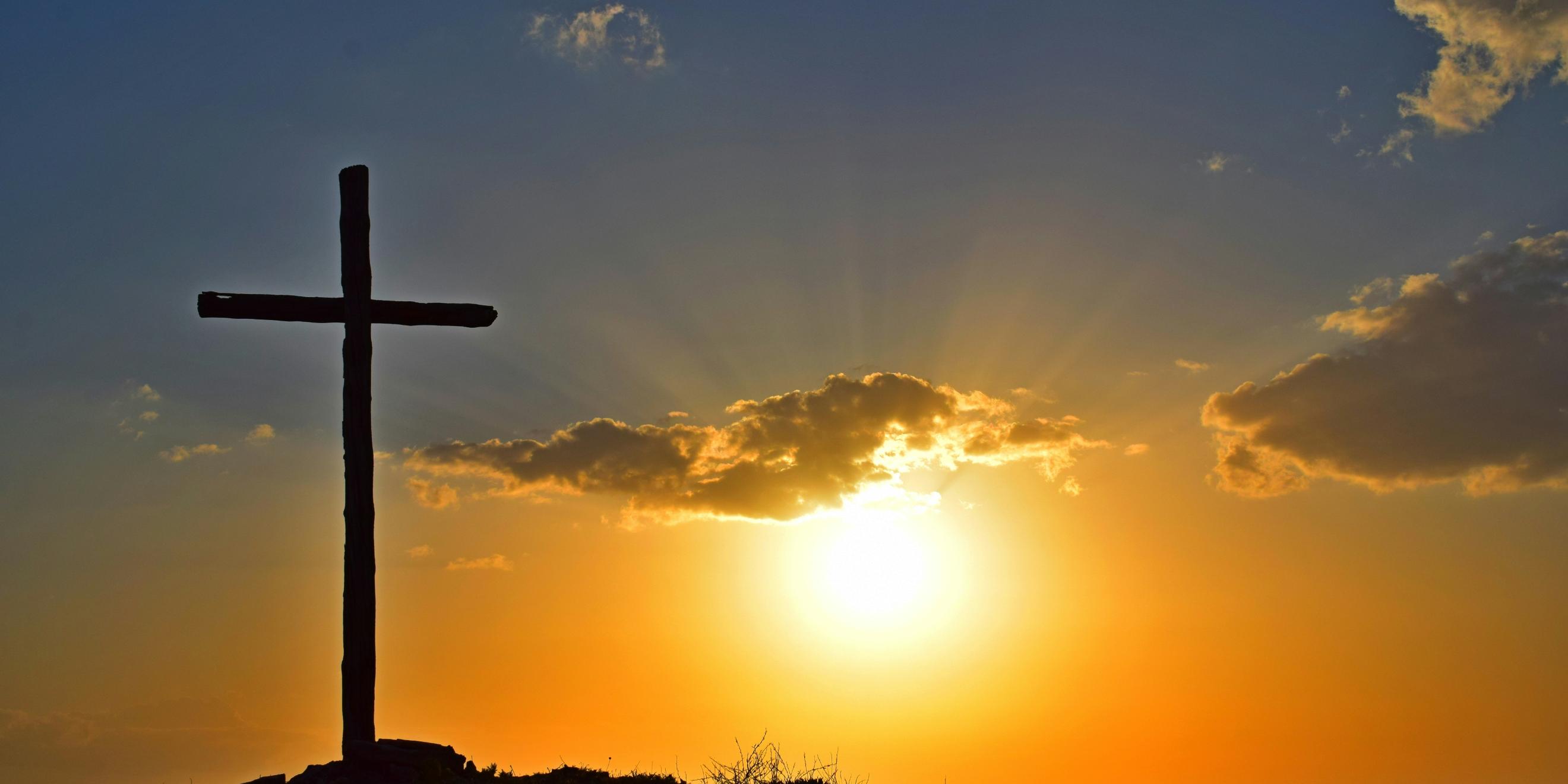 Auf einem Berg steht ein großes Kreuz, dahinter geht die Sonne unter