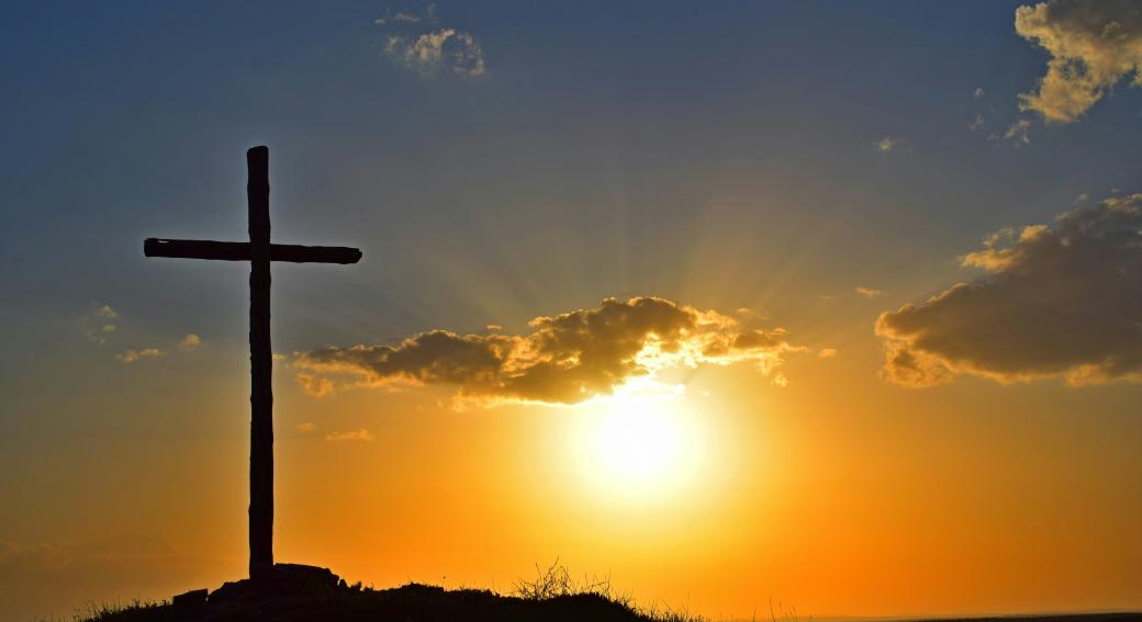 Auf einem Berg steht ein großes Kreuz, dahinter geht die Sonne unter