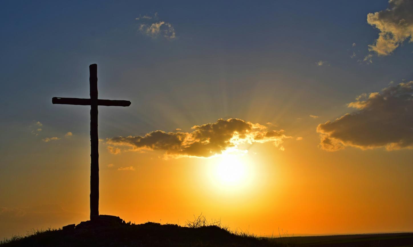 Auf einem Berg steht ein großes Kreuz, dahinter geht die Sonne unter