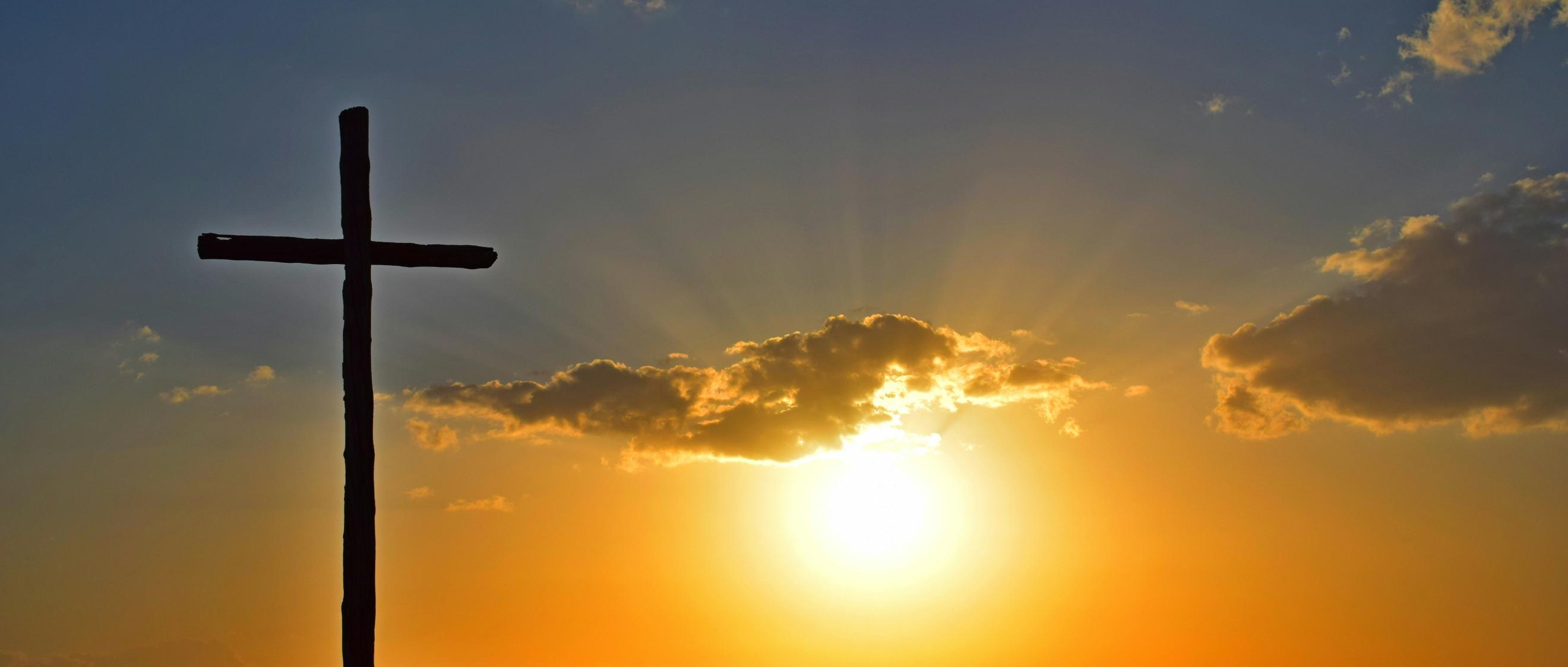 Auf einem Berg steht ein großes Kreuz, dahinter geht die Sonne unter